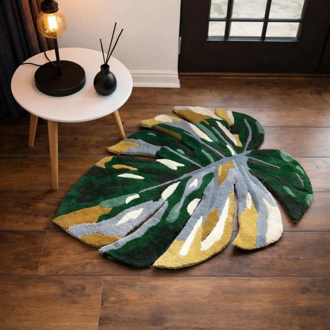 An angled view of the Monstera Tropical Rug displayed on a dark wood floor near a small wooden side table with a lit lamp. The sunlight highlights the thick, tufted texture and the intricate cut-out edges of the green, yellow, and grey leaf design.
