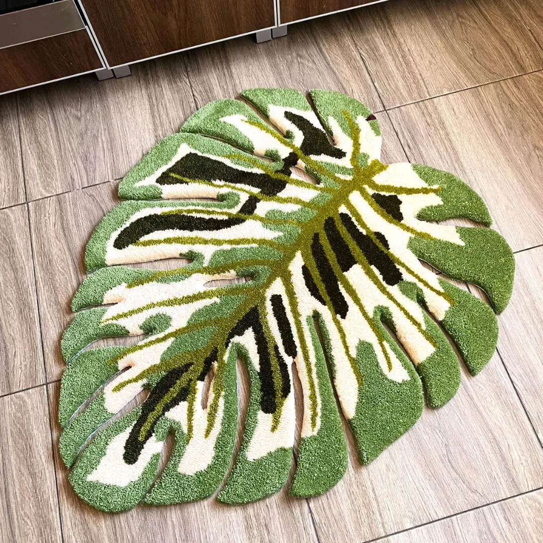 Full overhead view of the Verdant Monstera Rug laid flat on a wood-look tiled floor. The hand-tufted tropical leaf design features a white base with light green borders, a bright green central vein, and dark green shading.