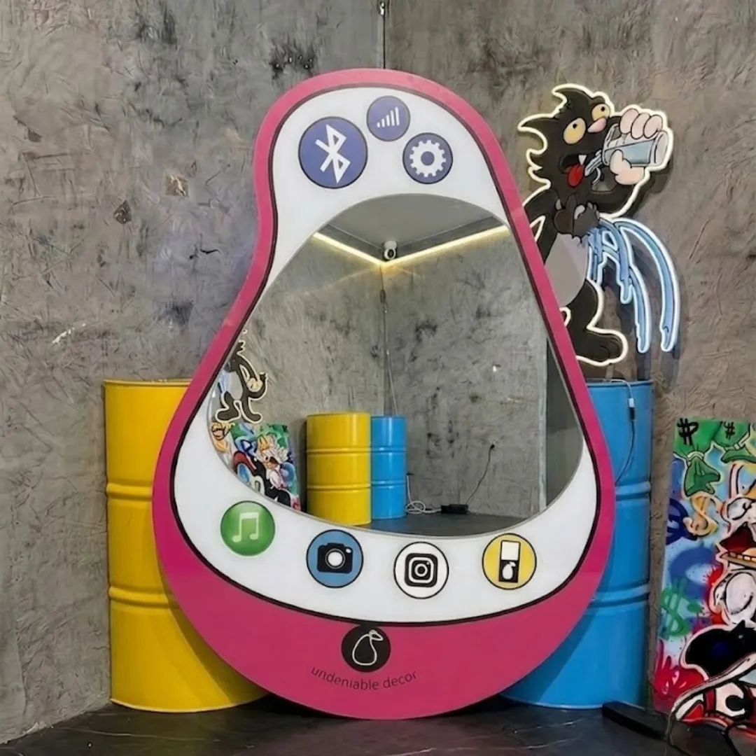 Interior styling view of the Aura Pear Phone Mirror displayed in a creative pop art studio space flanked by bright yellow and blue industrial barrels and glowing neon cartoon wall decor.