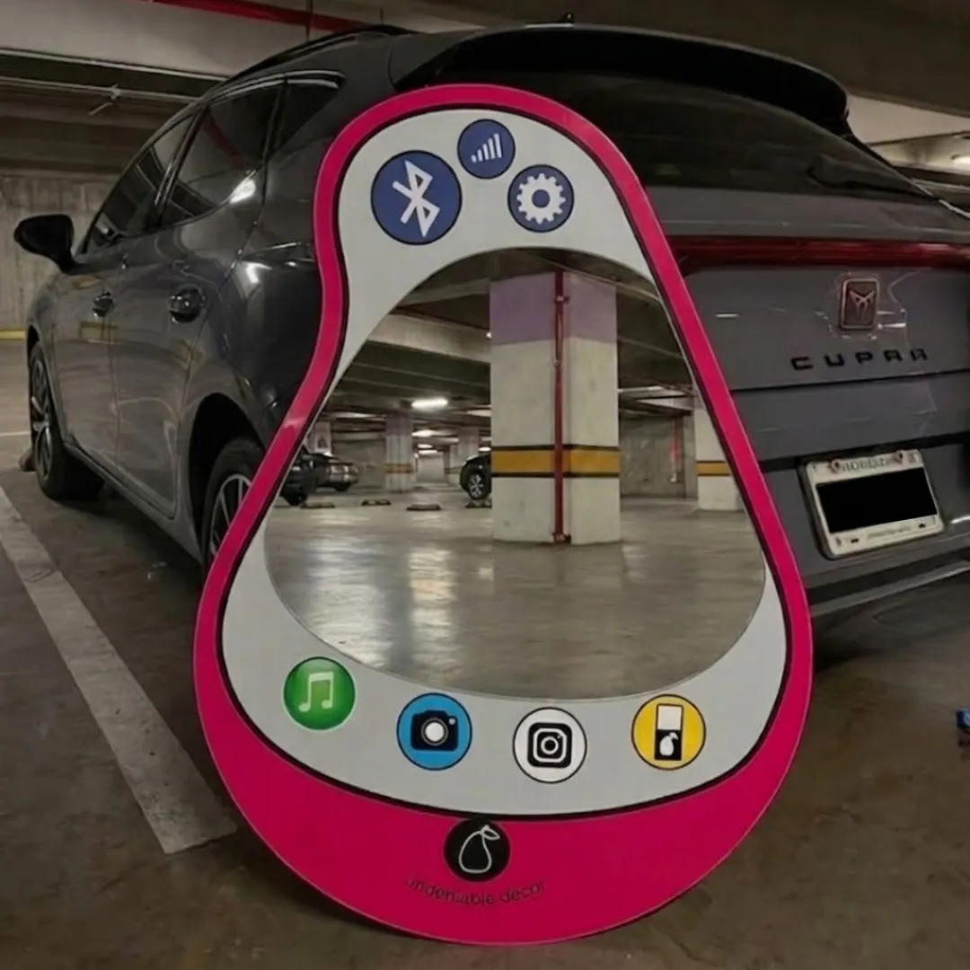Wide angle lifestyle shot of the hot pink Aura Pear Phone Mirror resting on the floor of a concrete underground parking garage, reflecting the interior columns and a grey vehicle.