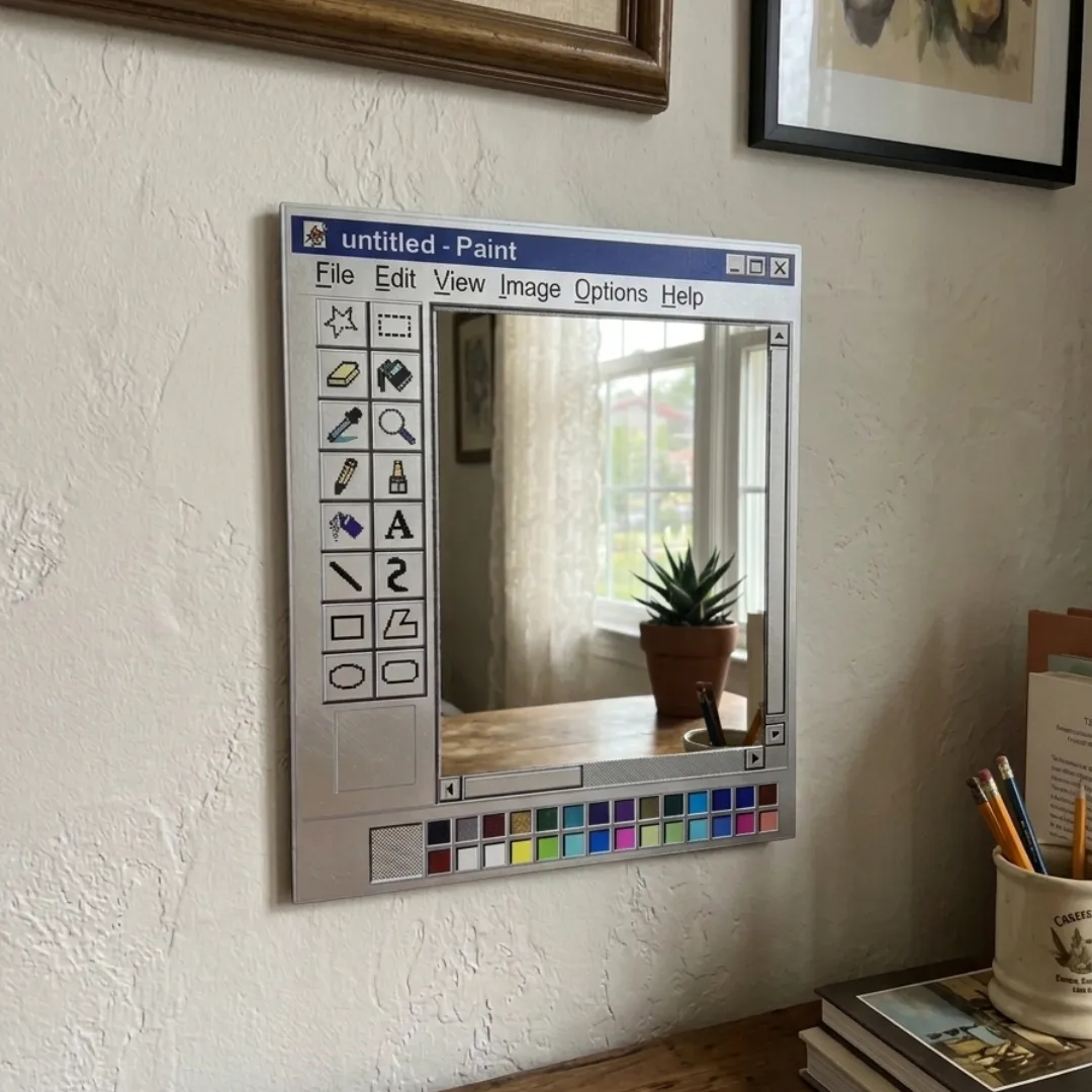An interior lifestyle view of a Paint Window mirror mounted on a white wall above a wooden desk, reflecting a window with a potted succulent, surrounded by a pencil holder and stacked books.