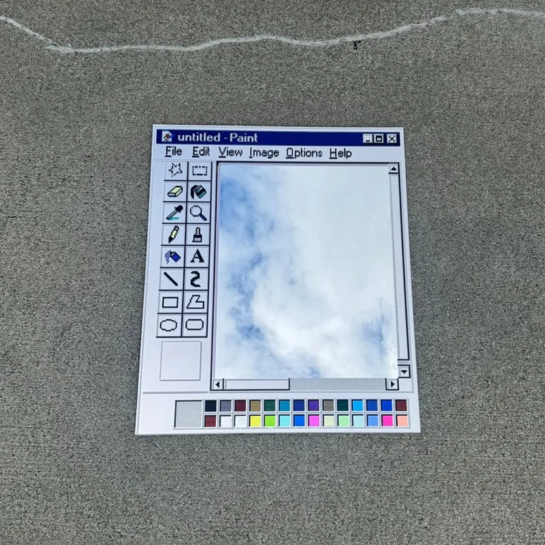 A high-angle close-up product shot of a Paint Window mirror isolated on a grey concrete surface, featuring a classic vintage computer paint software interface frame and a clear reflective glass mirror center.