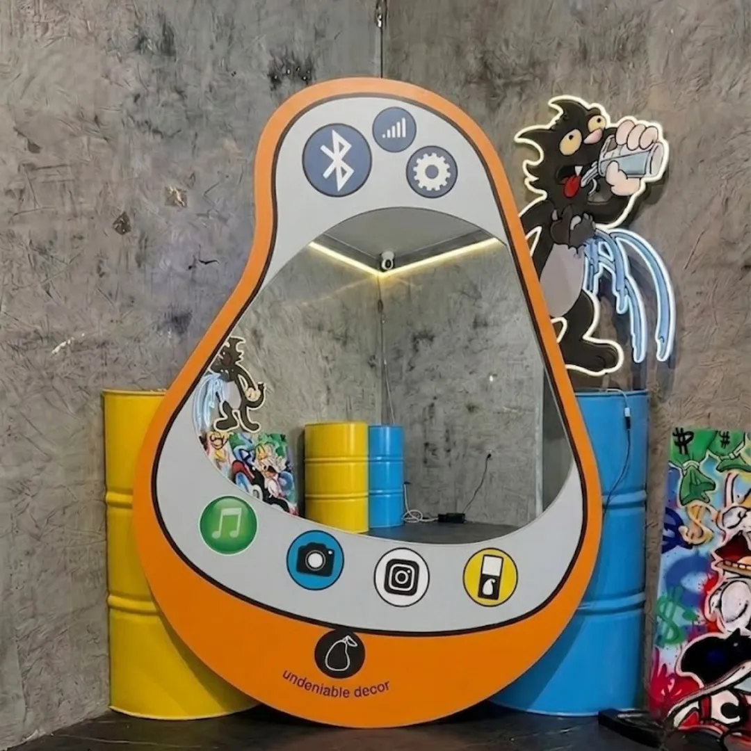 Interior styling view of the Pear Pop Phone Mirror displayed in a creative pop art studio space flanked by bright yellow and blue industrial barrels and glowing neon cartoon wall decor.