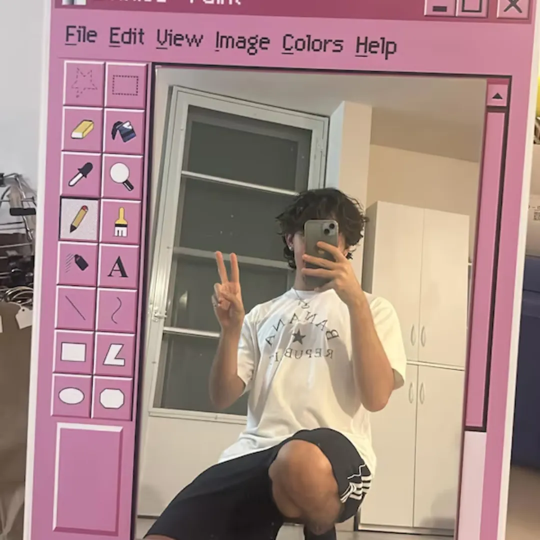 A person posing in front of a pink Pixel Window mirror, designed to look like a vintage computer paint application interface, placed in a bedroom setting.
