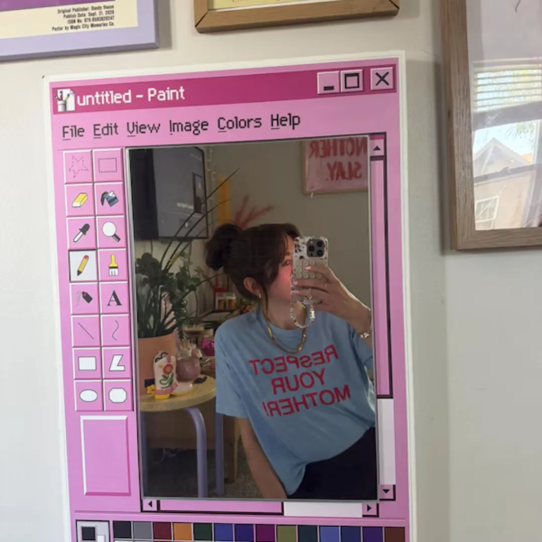 A person taking a selfie in a pink Pixel Window mirror, designed to look like a vintage computer paint application interface, mounted on a wall in a bedroom.