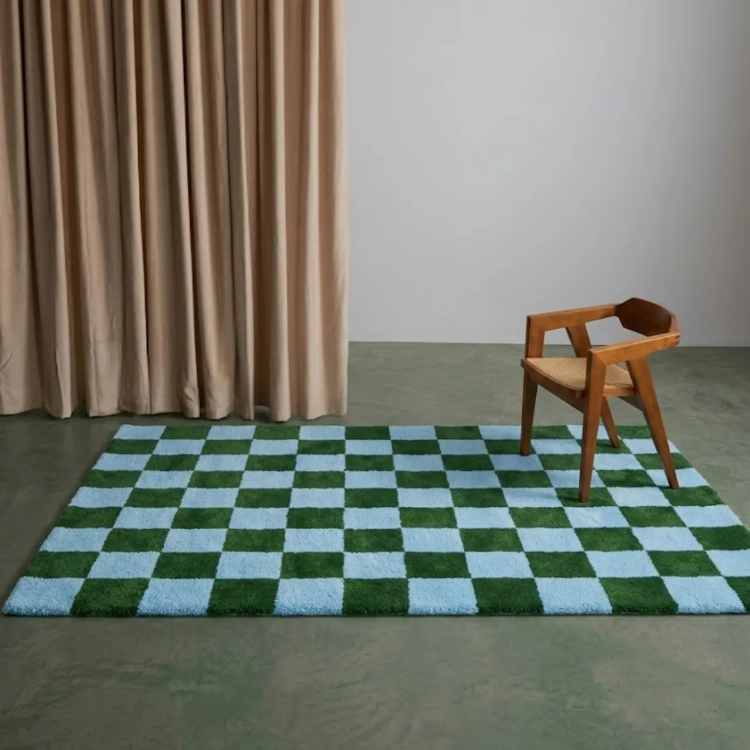 An interior lifestyle shot of the rectangular Sage Check rug styled in a minimalist room with a wooden chair, light-colored curtains, and a clean, neutral floor.