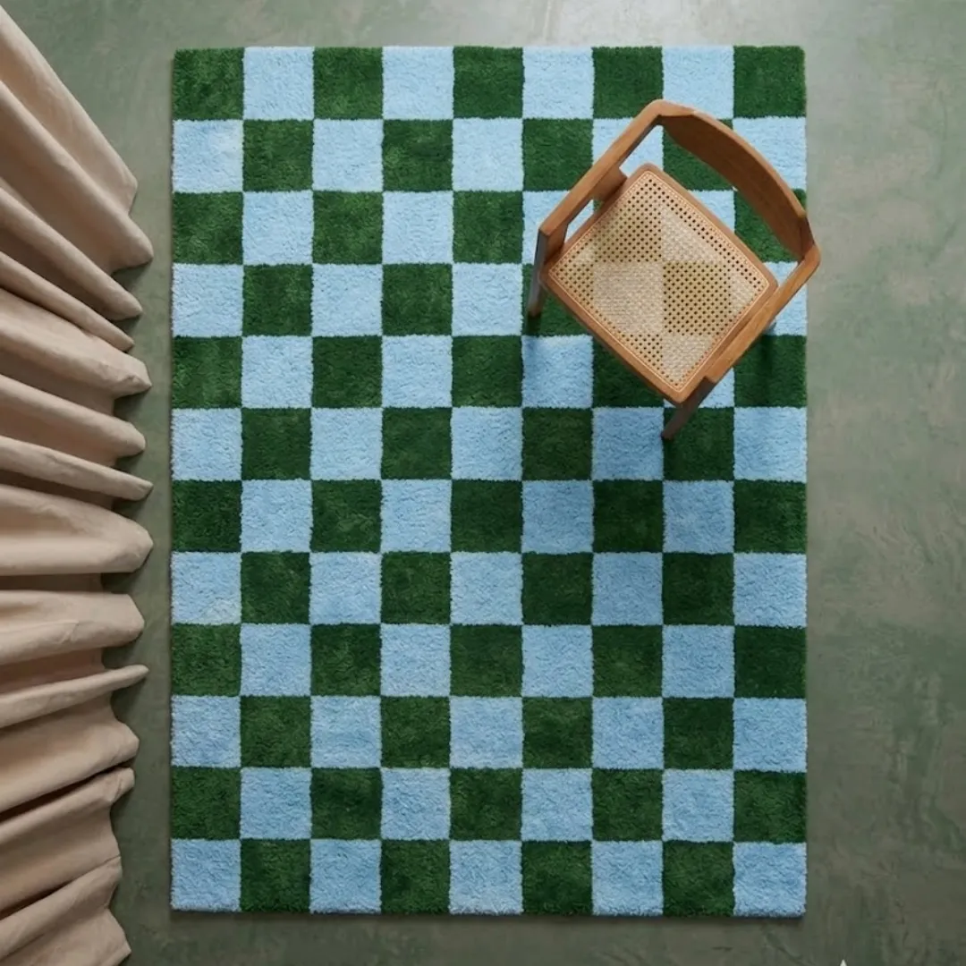 A top-down perspective interior shot of the Sage Check rug positioned on a smooth floor near a wooden chair and soft drapery, highlighting the vivid checkerboard design.