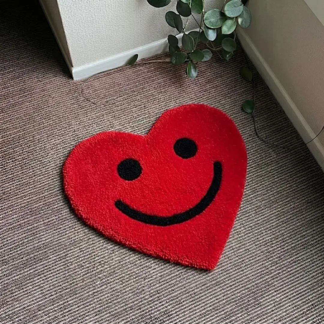 amora smile rug top view Amora Smile handmade tufted area rug shaped like a red heart with a black smiley face expression, displayed flat on a carpeted floor.