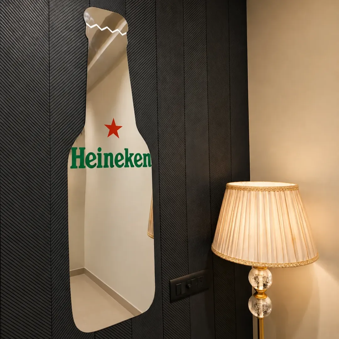 Bottle Shape decorative wall mirror featuring a custom Heineken beverage bottle design with a red star and green lettering, mounted on a dark textured wall next to a table lamp.