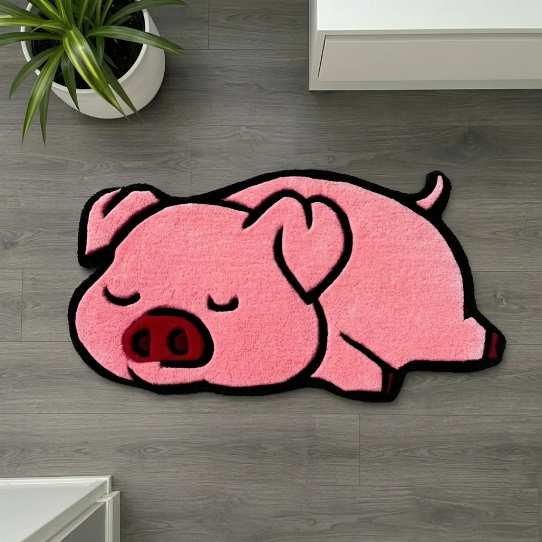 poppy snore rug overhead view Poppy Snore handmade tufted area rug featuring a cute sleeping pink pig shape with a dark red snout, displayed flat on a dark wooden floor next to a potted plant.