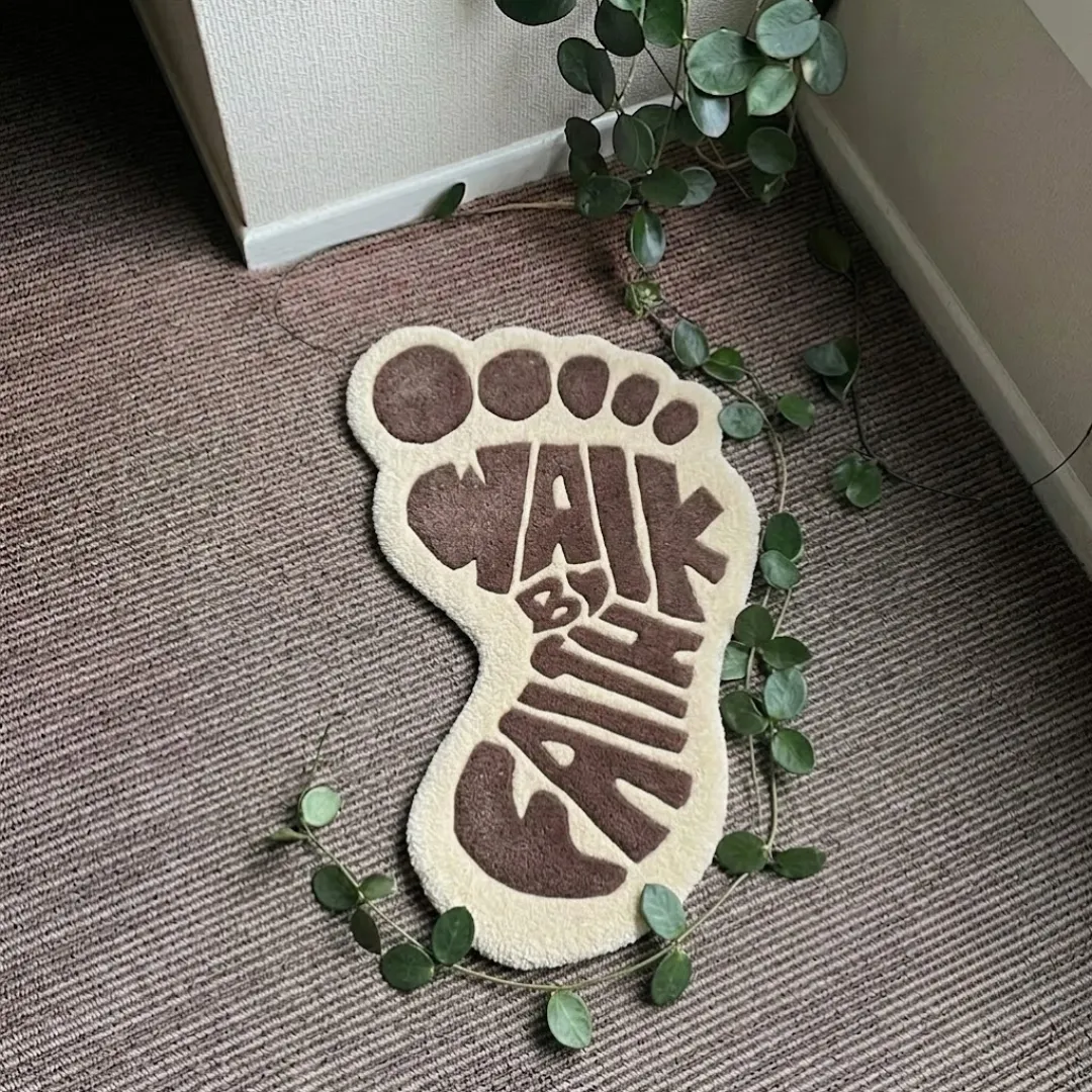 walk by faith rug top view Walk By Faith handmade tufted area rug shaped like a foot with brown typography and cream background, displayed flat on a carpeted floor.