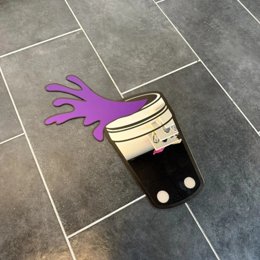 water spill mirror top view Water Spill shaped decorative wall mirror featuring a cup with a purple liquid splash, displayed flat on a dark tiled floor.